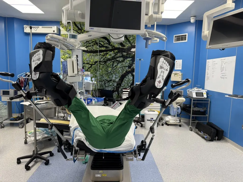 An operating table with leg supports, demonstrated by a person in green scrubs lying with legs raised.