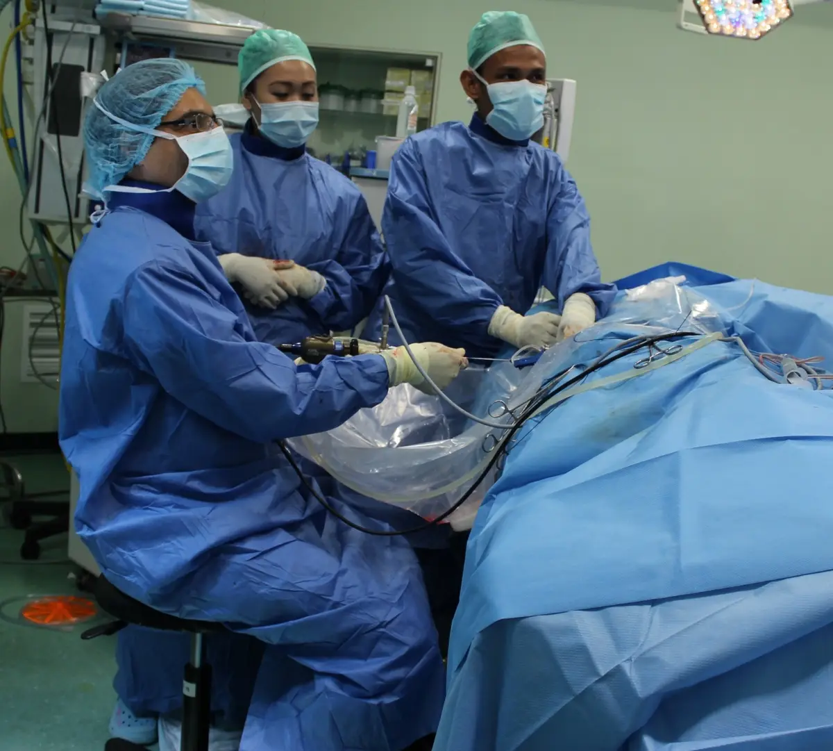 A surgeon in blue gown sitting on a stool while operating through the side of a draped patient, with two assistants standing behind him.
